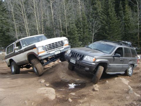1979 Ford Bronco - Two off-road vehicles, a white Ford Bronco and a gray Jeep Grand Cherokee, are navigating a rocky, dirt trail in a wooded area. The Bronco…