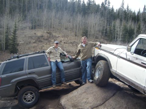 1979 Ford Bronco - Two men pose with their off-road vehicles, a dark gray SUV and a white pickup truck, positioned on large rocks in a wooded area.
