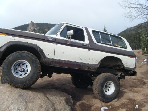 1979 Ford Bronco - A lifted 1979 Ford Bronco is shown from a low angle, driving on a rocky, off-road trail with a wooded hillside in the background.