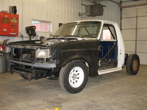 1994 Ford F-150 - Three-quarter front view of a white Ford pickup in a shop with the grille, hood and doors removed, exposing a black primered fender, bare r…