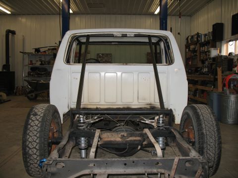 1994 Ford F-150 - Rear view of a white pickup truck chassis with a roll cage installed. The truck is in a workshop with various tools and equipment visible i…