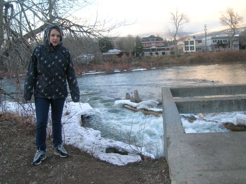 1996 Ford Bronco - A young woman stands on a dirt path next to a river with snow and ice along the bank. Buildings and trees are visible on the opposite bank…