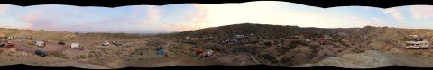 1984 Ford Bronco - A panoramic view of a desert campsite at dusk with multiple RVs, trailers, tents, and vehicles parked in a dry, hilly landscape.