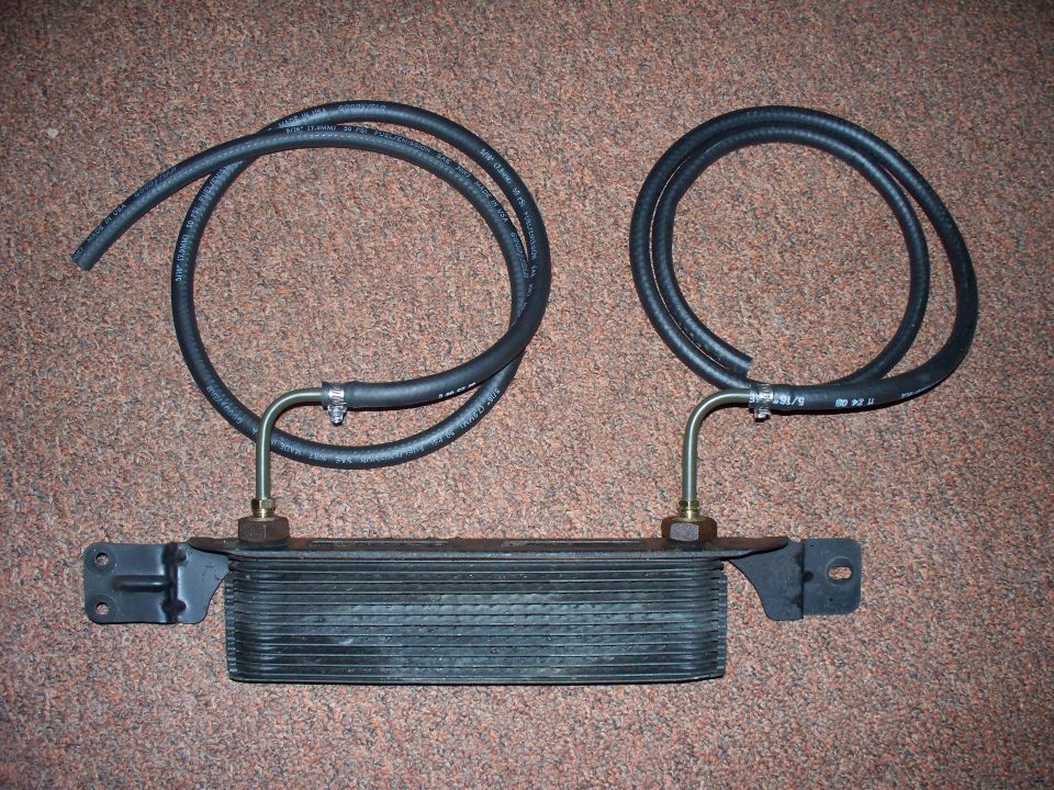 Finned oil/transmission cooler with brackets, brass fittings, and two coiled black rubber hoses on carpet