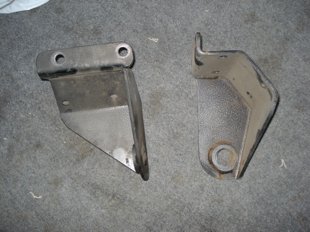 Two metal brackets on a carpeted surface. One bracket has two holes on top and a triangular plate. The other bracket is L-shaped with a circular hole at the bottom