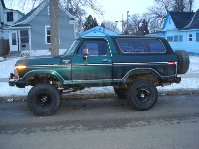 Lifted dark green Ford Bronco with big tires and rear spare, side view on a snowy street