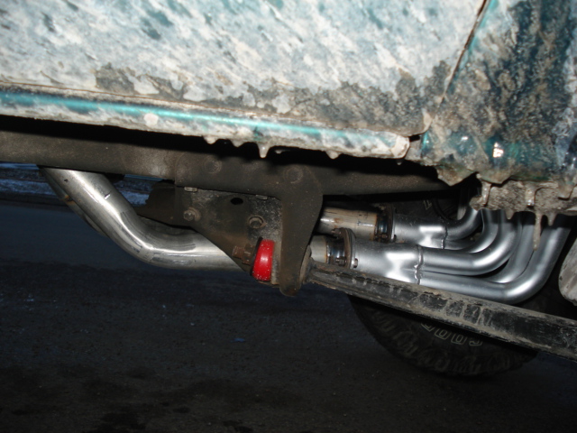 Under-body view of a green vehicle showing chrome headers, exhaust pipe, and red body mount bushing