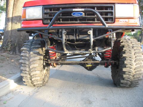 1987 Ford Bronco - Low front view of a lifted red Ford 4x4 parked at a curb, showing the blue Ford oval on the grille, a 4x4 fender badge and a black tube bum…