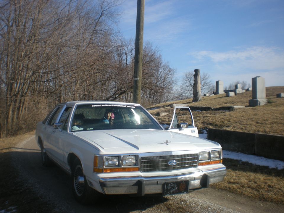 White sedan parked on a dirt road next to a cemetery, driver's door open