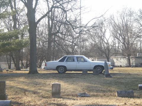1988 Ford Crown Victoria LTD - A white 1988 Ford Crown Victoria LTD sedan is seen parked in a cemetery with bare trees and gravestones visible.