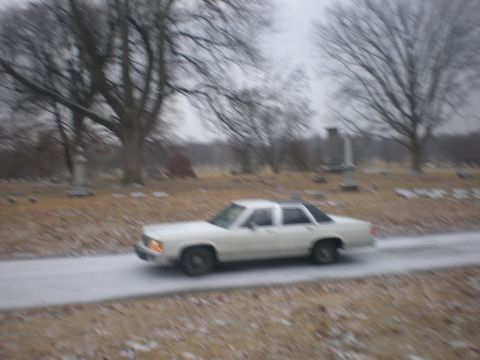 1988 Ford Crown Victoria LTD - A white sedan drives through a cemetery with bare trees and gravestones visible.