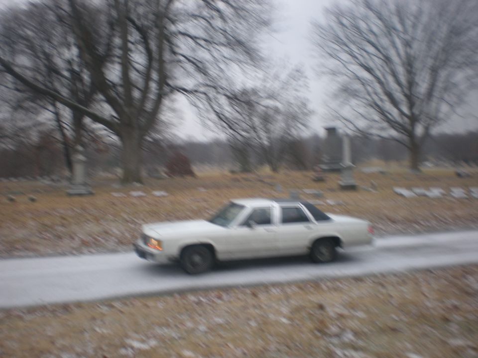 White boxy Ford sedan driving through a cemetery lane in winter, motion-blurred side view