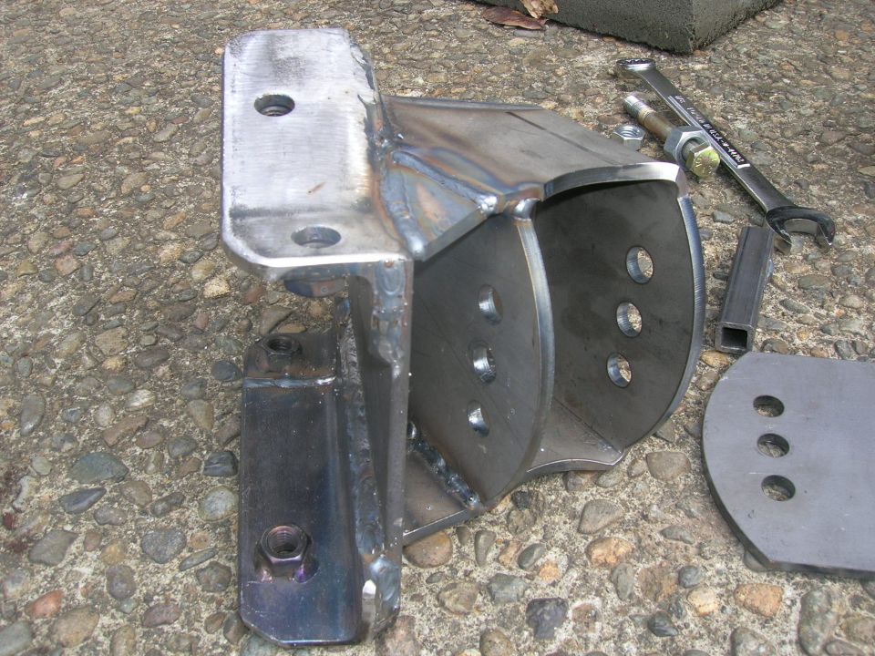 Custom metal bracket with multiple mounting holes, next to a wrench and bolts on a gravel surface