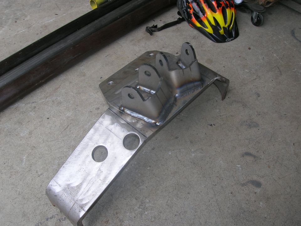 Welded metal bracket with two mounting holes and two upper attachment points, sitting on concrete floor