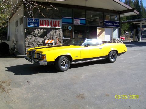 1973 Mercury Cougar - A bright yellow 1973 Mercury Cougar convertible, a black racing stripe, is parked outside a Cedar Glen Auto Care service station.