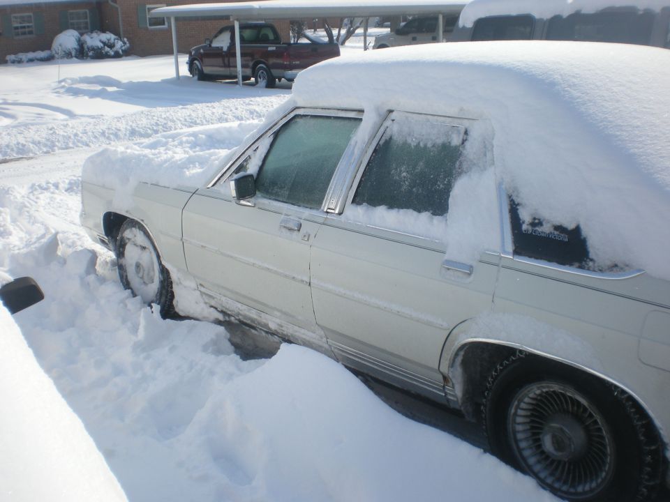 1988 Ford Crown Victoria LTD covered in snow, parked in a driveway. Another truck is visible under a carport