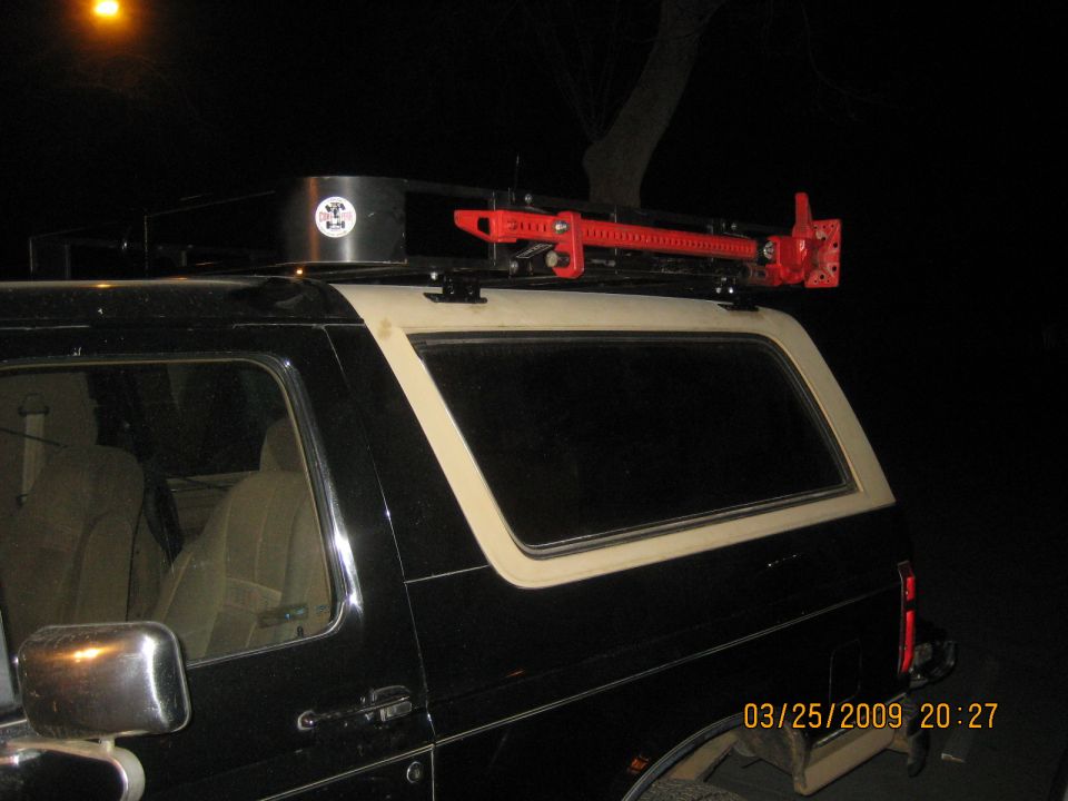 Black Ford Bronco with white top, roof rack and red Hi-Lift jack at night