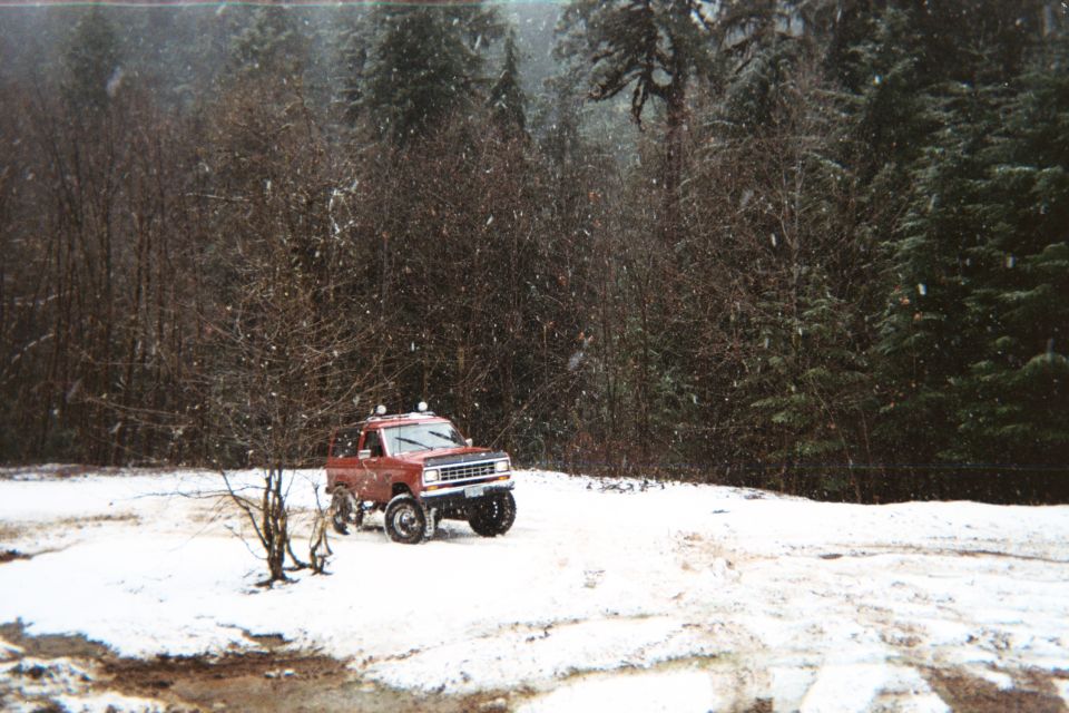 Lifted red Ford Bronco II parked on snowy ground with falling snow and forest behind