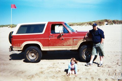 1985 Ford Bronco - A man and a young girl pose with a red Ford Bronco on a sandy beach under a clear blue sky.