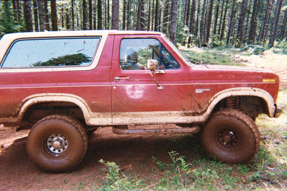 Side view of a red 1985 Ford Bronco covered in mud, driving on a dirt path in a forest