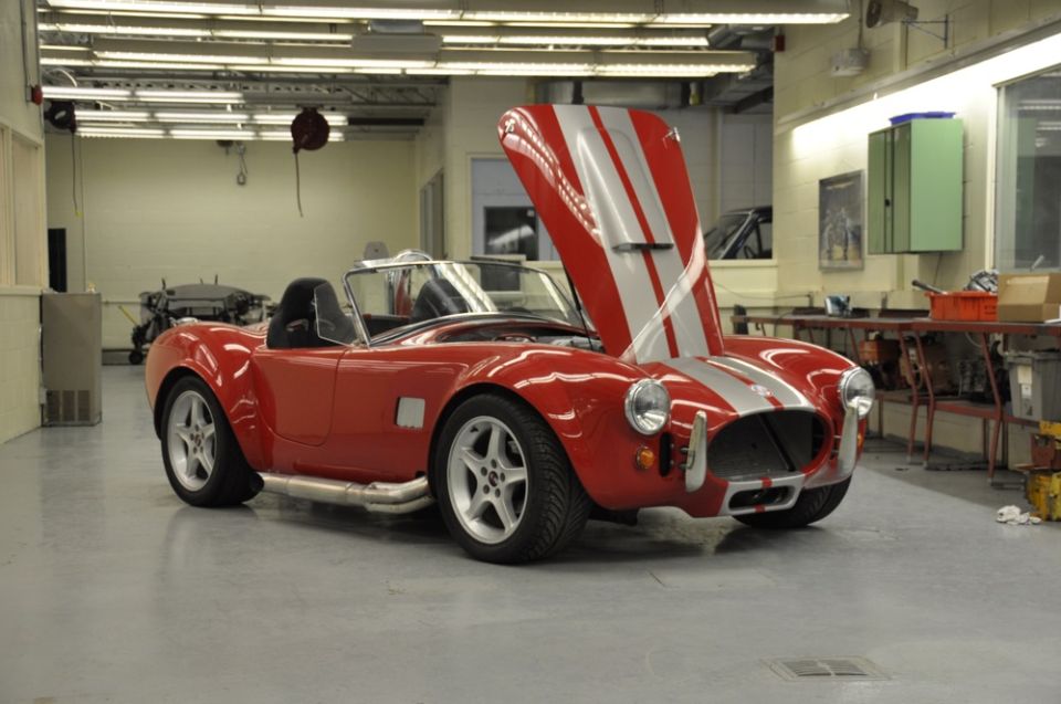 Red convertible sports car with silver stripes on hood, hood is open. Car is in a garage with tools and equipment in the background