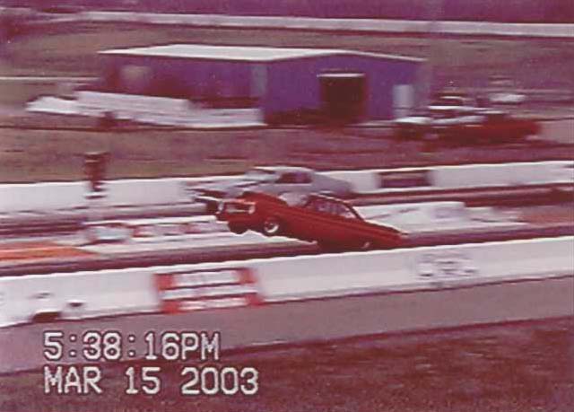 Red drag car racing on a track, another car visible in background. Timestamp reads 5:38:16PM MAR 15 2003