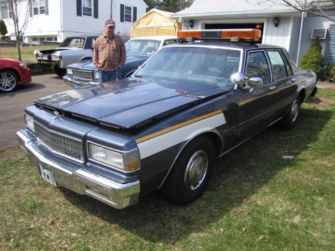 1991 Ford LTD Crown Victoria - This 1980s police car has its hood up and is parked on a grassy lawn.