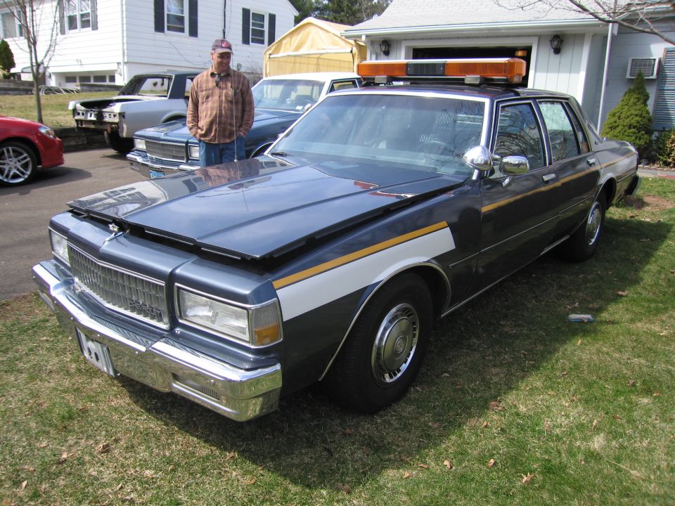1980s police car with light bar, hood up, parked on grass. Another car is behind it