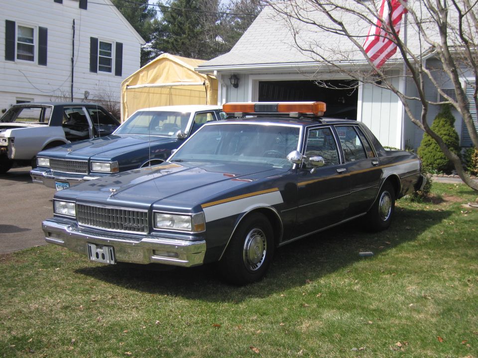 Dark blue police car with light bar, parked in front of a garage. Another blue car and a silver car are behind it