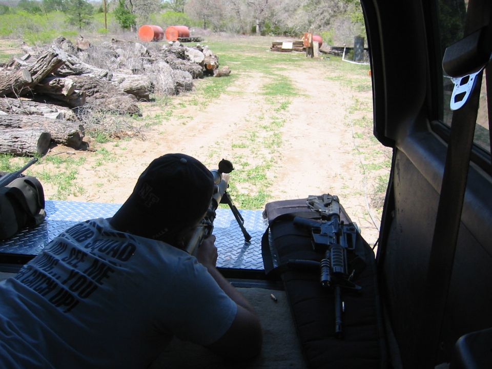 Man aiming rifle from inside vehicle, with another rifle on a bag beside him