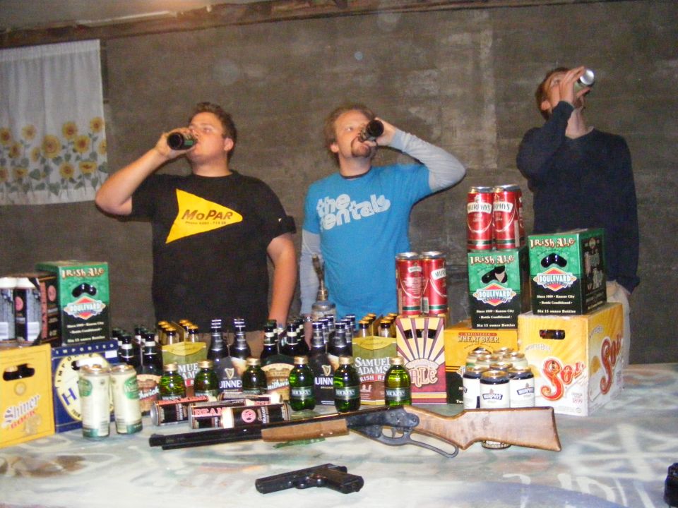 Three guys drinking beer and holding guns on a table covered in bottles and cans