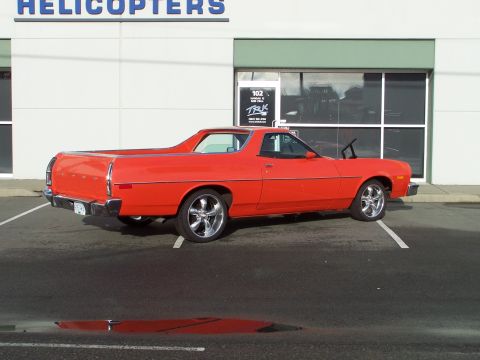 1973 Ford Ranchero - Driver-side rear three-quarter view of an orange-red Ford Ranchero coupe-utility on polished five-spoke wheels, parked in an empty lot outs…
