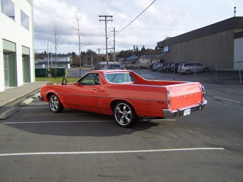 1973 Ford Ranchero - Driver-side rear three-quarter view of an orange-red Ford Ranchero coupe-utility parked in a commercial lot, showing the long body-color ca…