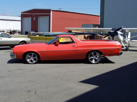 1973 Ford Ranchero - Full driver-side profile of a lowered orange-red Ford Ranchero coupe-utility on polished five-spoke wheels, parked on airport tarmac with a…