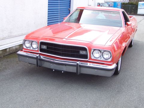 1973 Ford Ranchero - Three-quarter front view of a red Ford Ranchero coupe utility parked on asphalt beside a white building with blue roll-up doors. Quad round…