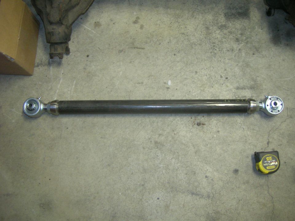Aftermarket suspension control arm with spherical bearings on each end, next to a tape measure