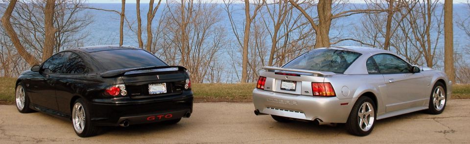 Black Pontiac GTO and silver Ford Mustang parked side-by-side with water and trees in the background
