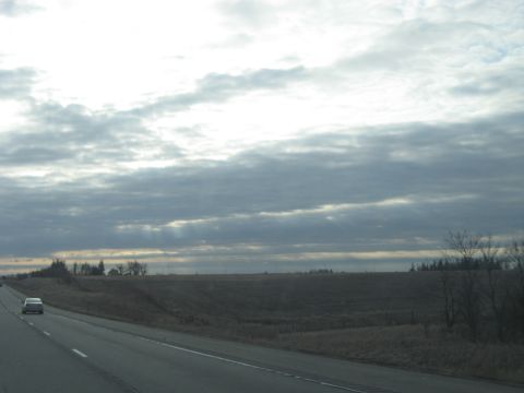 1978 Ford Bronco - A lone silver sedan travels down a highway, dwarfed by a vast, overcast sky and rural landscape.
