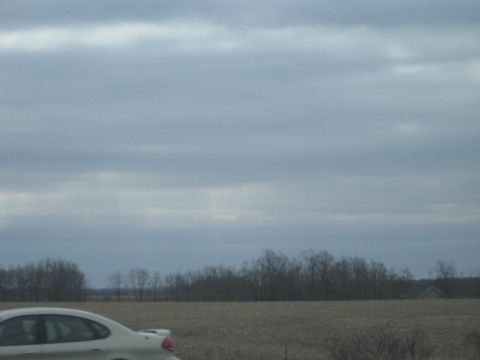 1978 Ford Bronco - A car travels down a road next to a barren field and trees under an overcast sky.