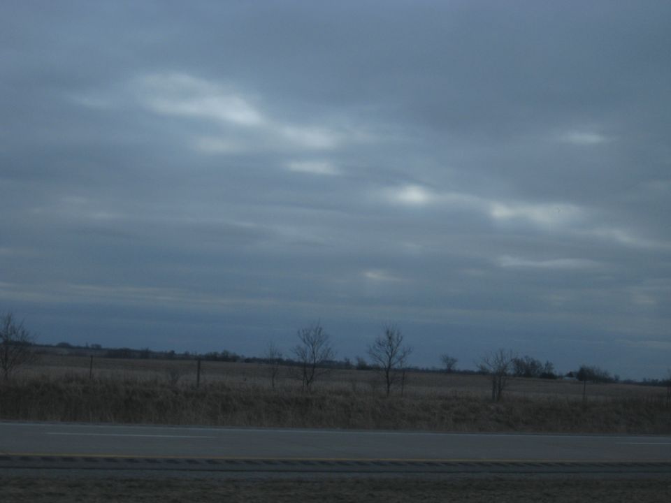 Overcast sky above a dry, grassy field with scattered bare trees and a highway in the foreground