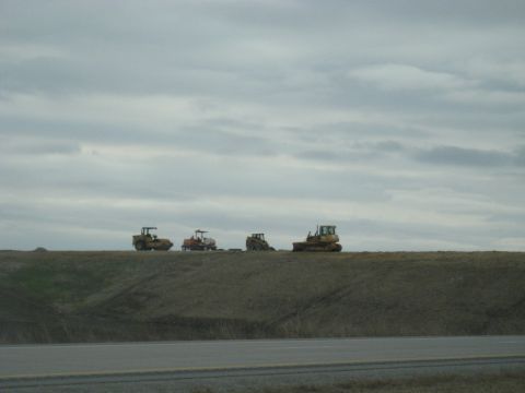 1978 Ford Bronco - A line of construction vehicles, including bulldozers and a roller, are parked on a ridge overlooking a highway.