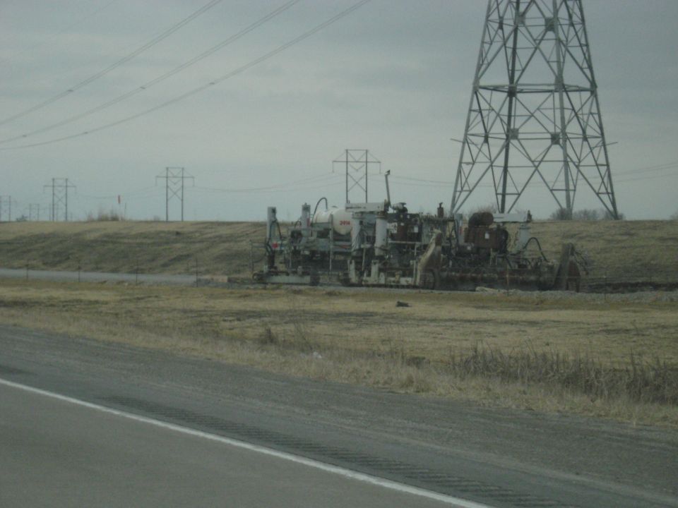 Road construction equipment parked in a field next to a highway with power lines in the background