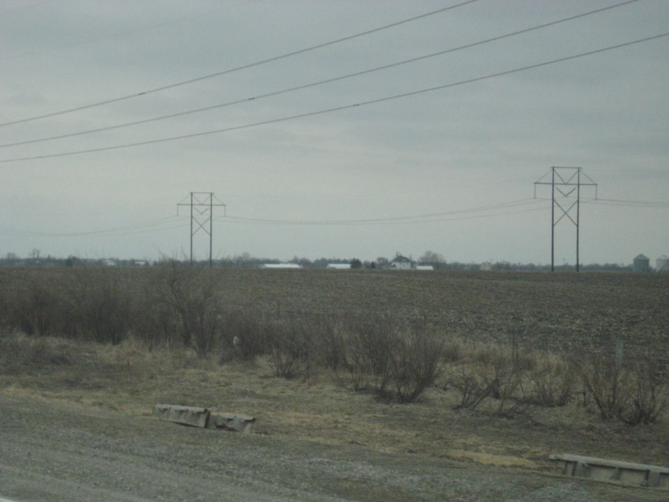 Power lines stretch across a cloudy sky over a barren field with distant farm buildings