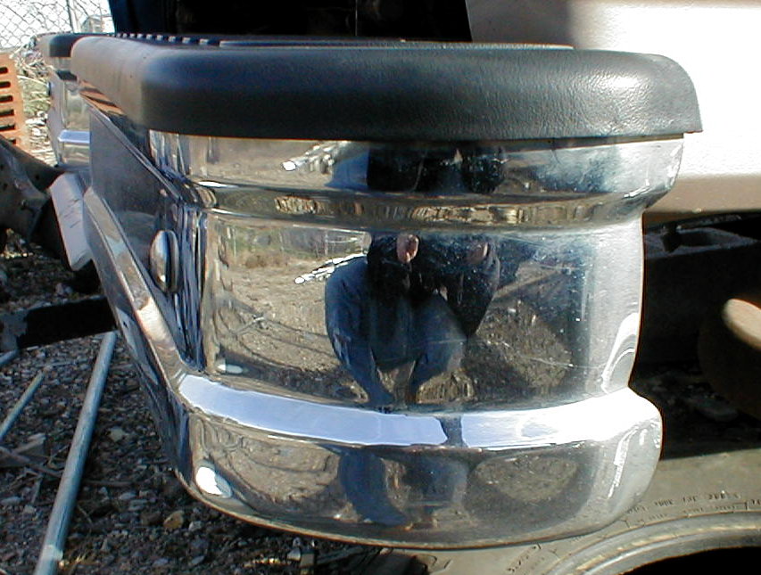 Chrome front bumper corner with black rubber cap reflecting a person and dirt lot
