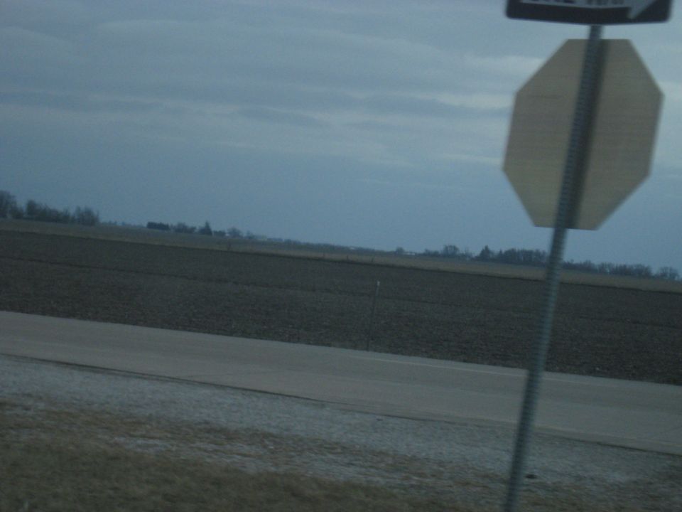 Roadside view of a field under a cloudy sky with a stop sign and a one-way sign