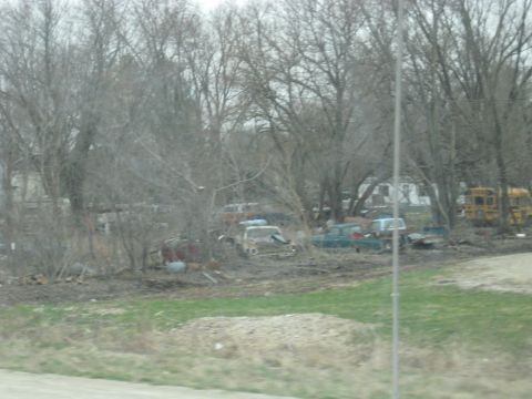 1978 Ford Bronco - A collection of abandoned vehicles sits in a lot surrounded by trees, with a yellow school bus visible on the right.