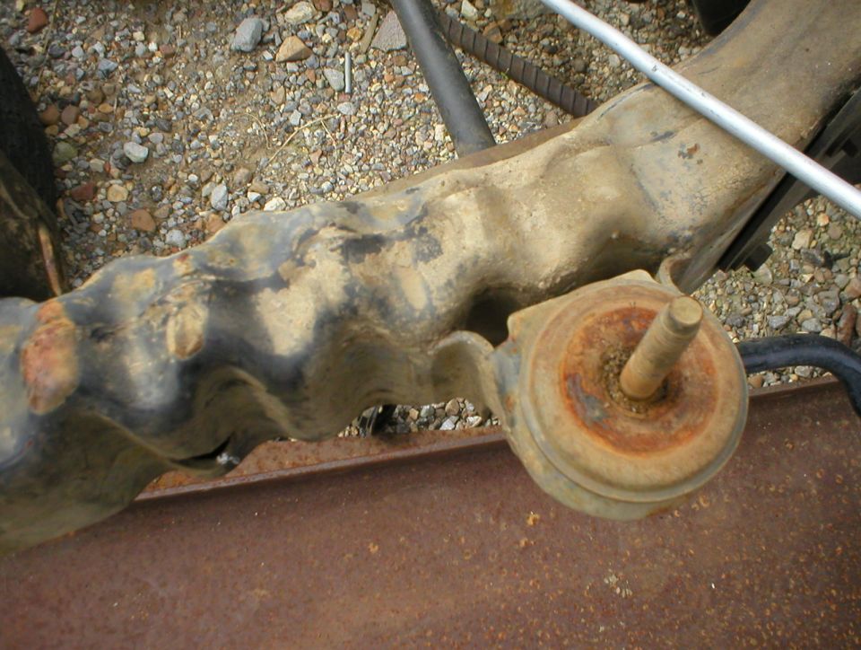 Rusty body mount stud and bracket on a weathered vehicle frame over gravel