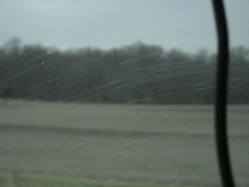 Overcast sky above a field and distant trees, viewed through a dirty windshield