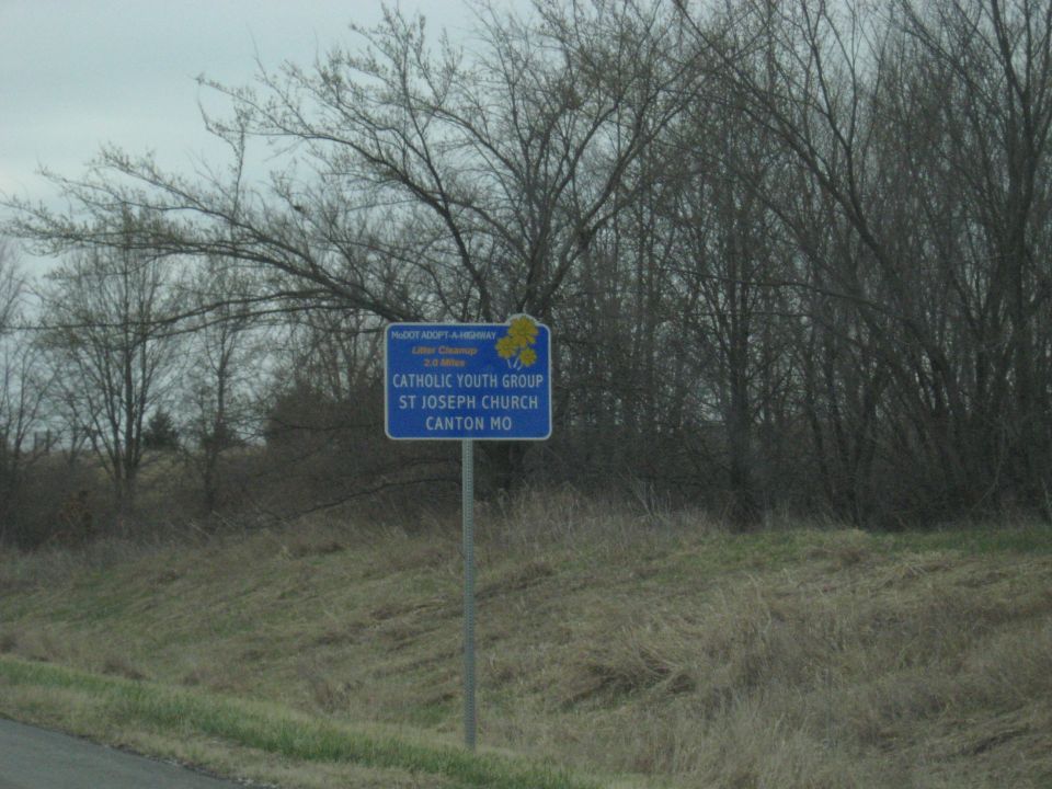 Blue sign with yellow flowers reads 'Adopt-A-Highway', 'Catholic Youth Group', 'St Joseph Church', 'Canton MO'. Bare trees and dry grass in background
