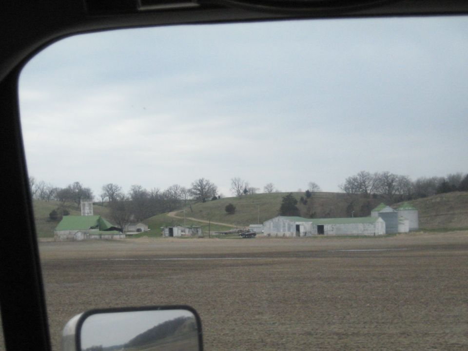 Farm buildings with green roofs and silos under a cloudy sky, viewed from a vehicle